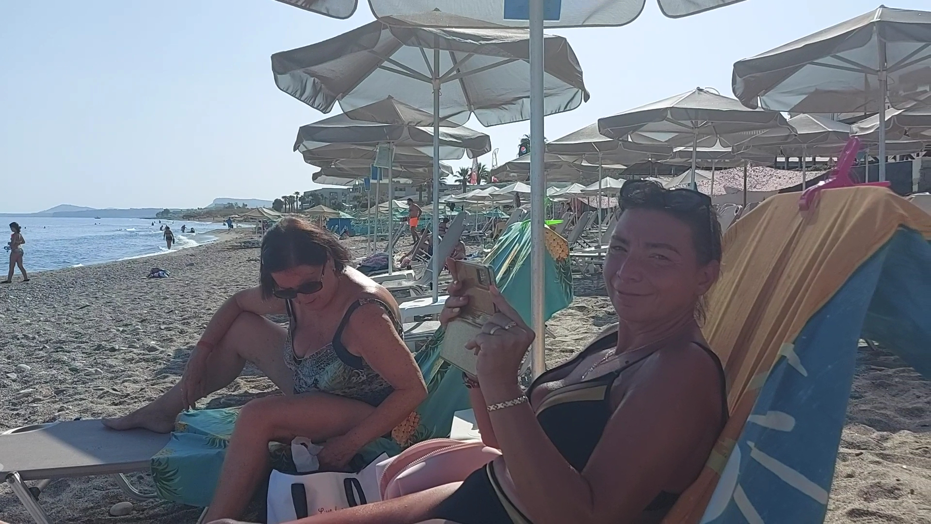 Zeus Hotels Ydoria Resort viesi atpūšas saulessargu ēnā oļu pludmalē Krētā, baudot skatu uz Vidusjūras dzidro ūdeni