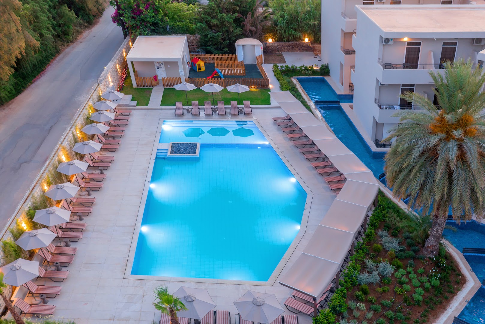 Zeus Hotels Ydoria Resort galvenais baseins vakarā no augšas Hersonisā, Krētā — apkārt sauļošanās krēsli, saulessargi un bērnu rotaļu laukums