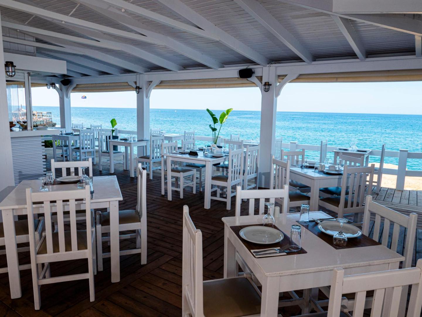 Plašs Porto Greco Village āra restorāns zem nojumes ar baltiem koka krēsliem un galdiem, skats uz Vidusjūru