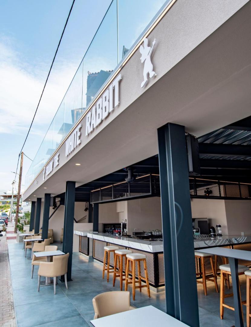 Porto Greco Village āra bārs White Rabbit ar modernu dizainu, augstajiem krēsliem un skatu uz ielu Hersonisas piekrastē