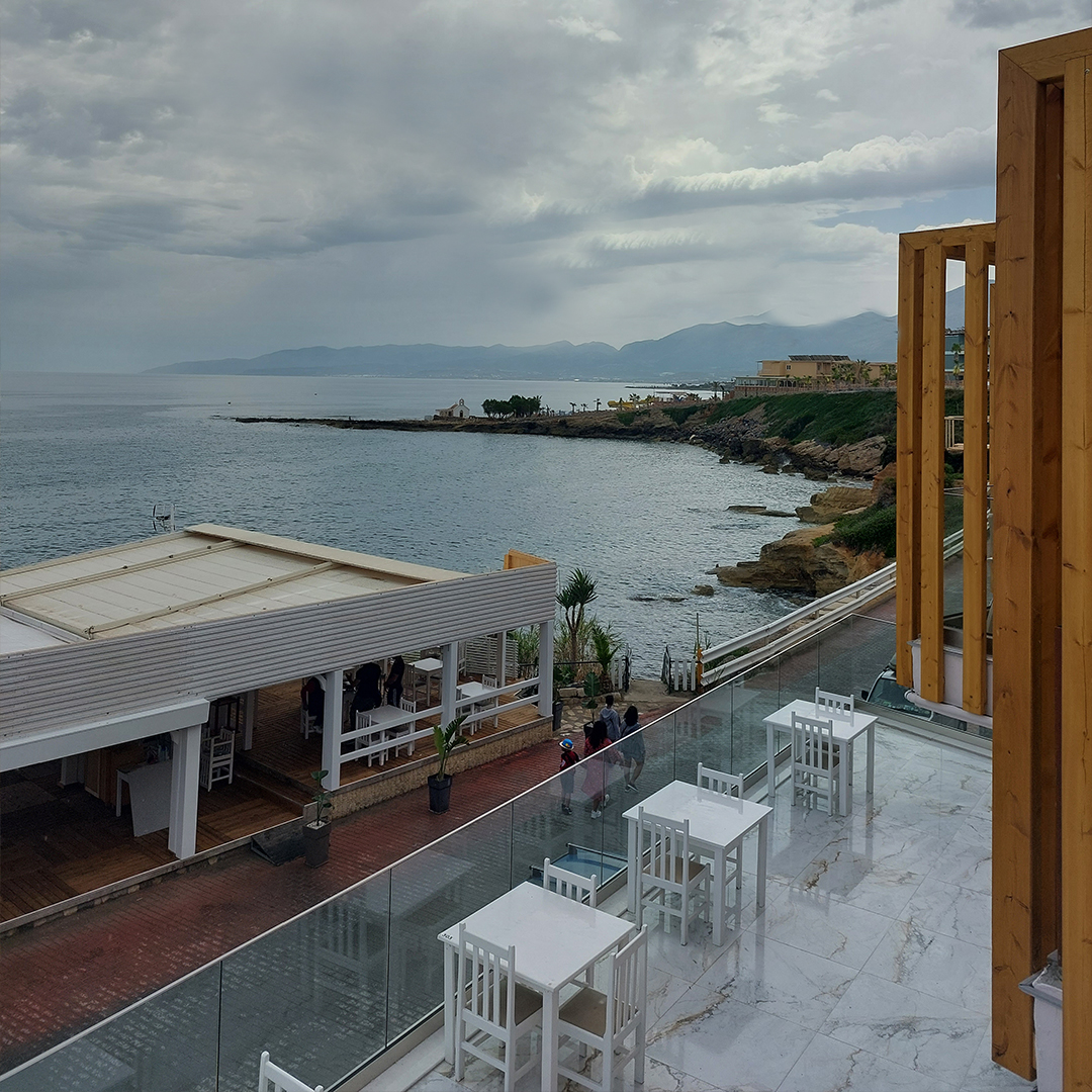 Porto Greco Village viesnīcas āra terase ar skatu uz jūru un piekrastes līniju Hersonisā, Krētā — balti galdiņi un marmora grīda