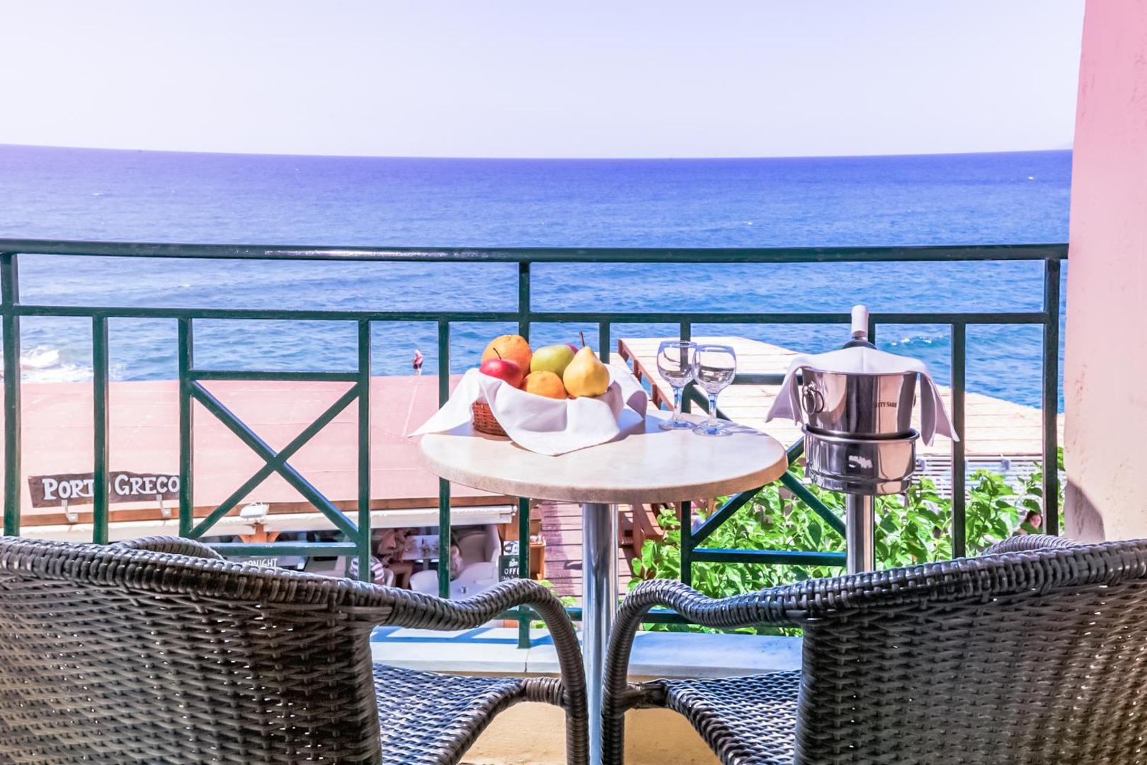 Porto Greco Village balkons ar pītiem krēsliem un skatu uz zilo jūru — galds ar augļiem un dzērieniem