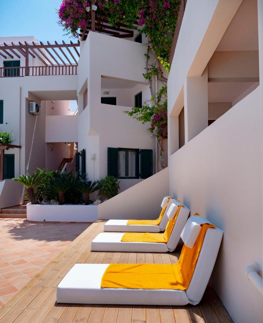 Porto Greco Village saulaina terase ar ērtiem sauļošanās krēsliem un koši dzelteniem dvieļiem pie viesnīcas baltajām ēkām