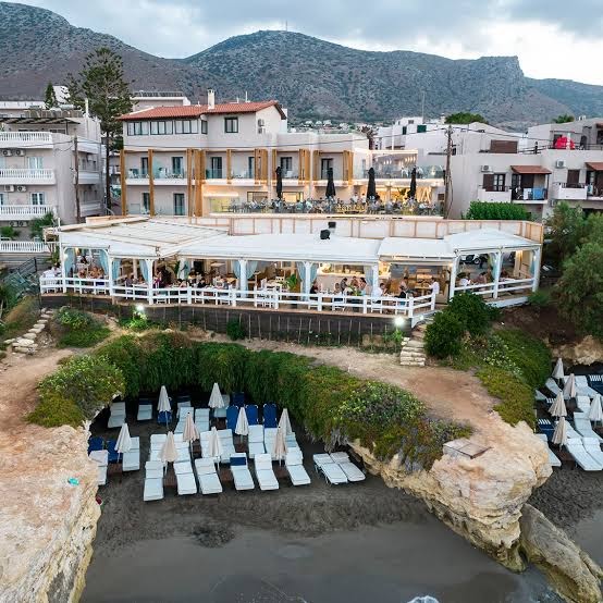 Porto Greco Village viesnīcas restorāna terase uz klints malas virs pludmales zonas ar sauļošanās krēsliem un saulessargiem vakarā