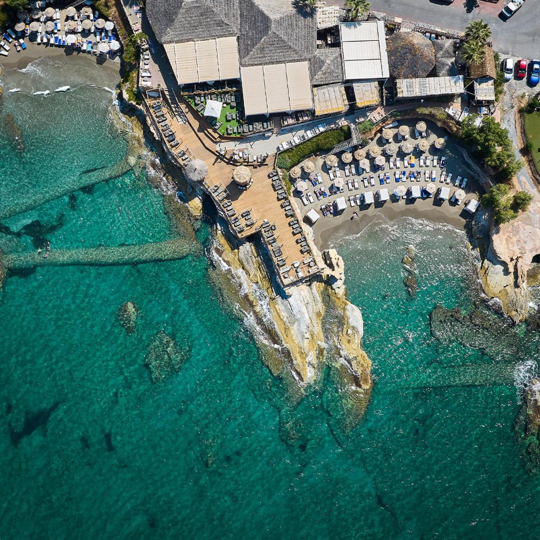 Porto Greco Village skats no putna lidojuma uz piekrastes restorānu un nelielu pludmales līci ar tirkīzzilu ūdeni Hersonisā, Krētā