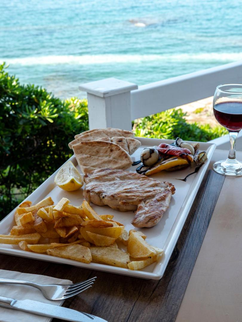Porto Greco Village restorāna piedāvājums — grilēta gaļa ar frī kartupeļiem, dārzeņiem un vīna glāzi uz jūras fona