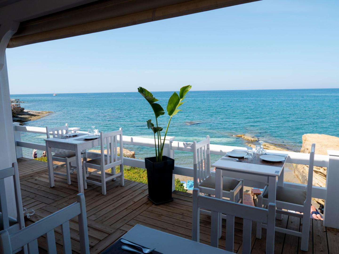 Porto Greco Village restorāna koka terase tieši pie jūras ar baltām mēbelēm un skatu uz dzidri zilo ūdeni Krētā
