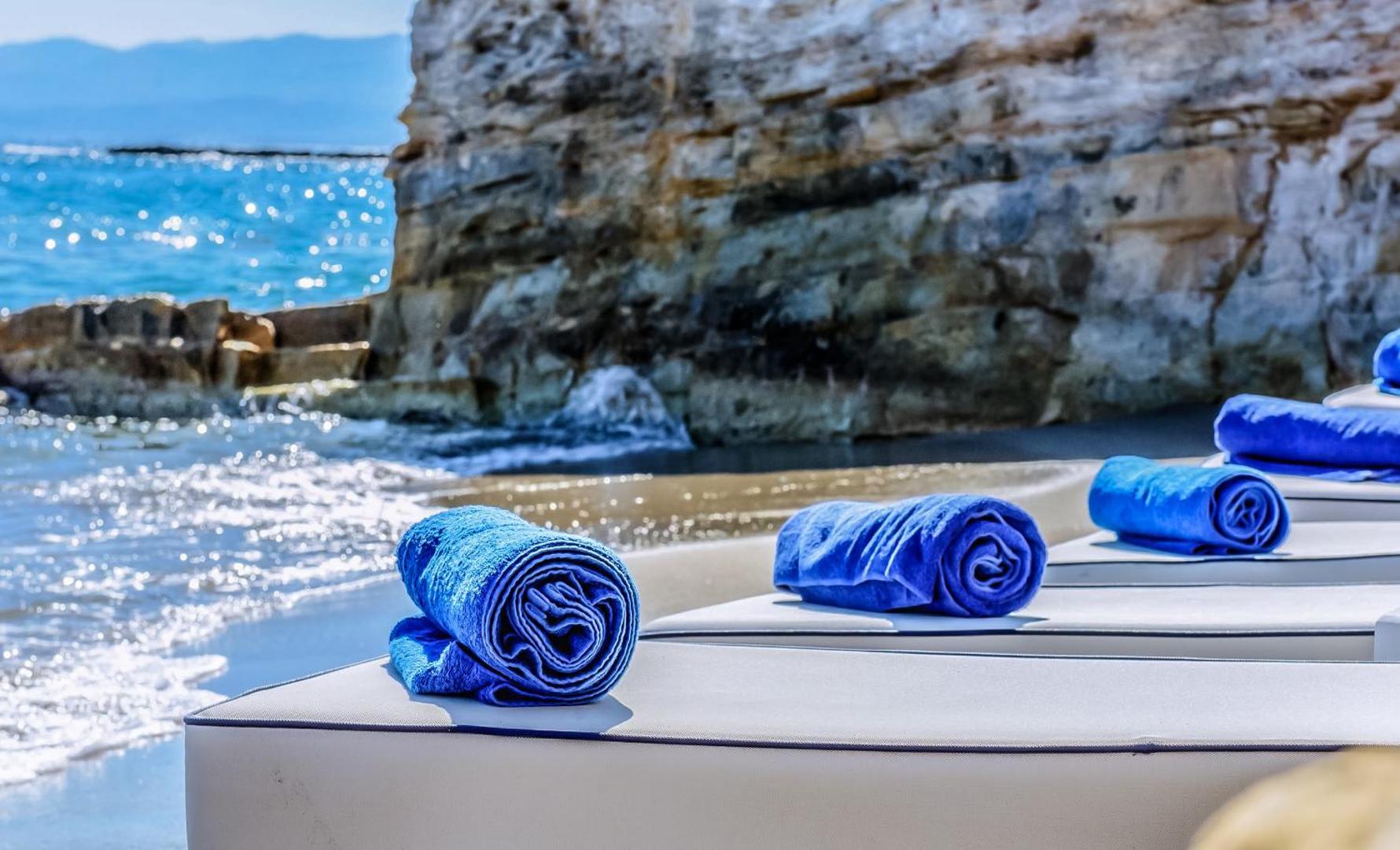 Porto Greco Village pludmales atpūtas zona ar mīkstiem sauļošanās krēsliem un ziliem dvieļiem pie klints sienas Hersonisā, Krētā