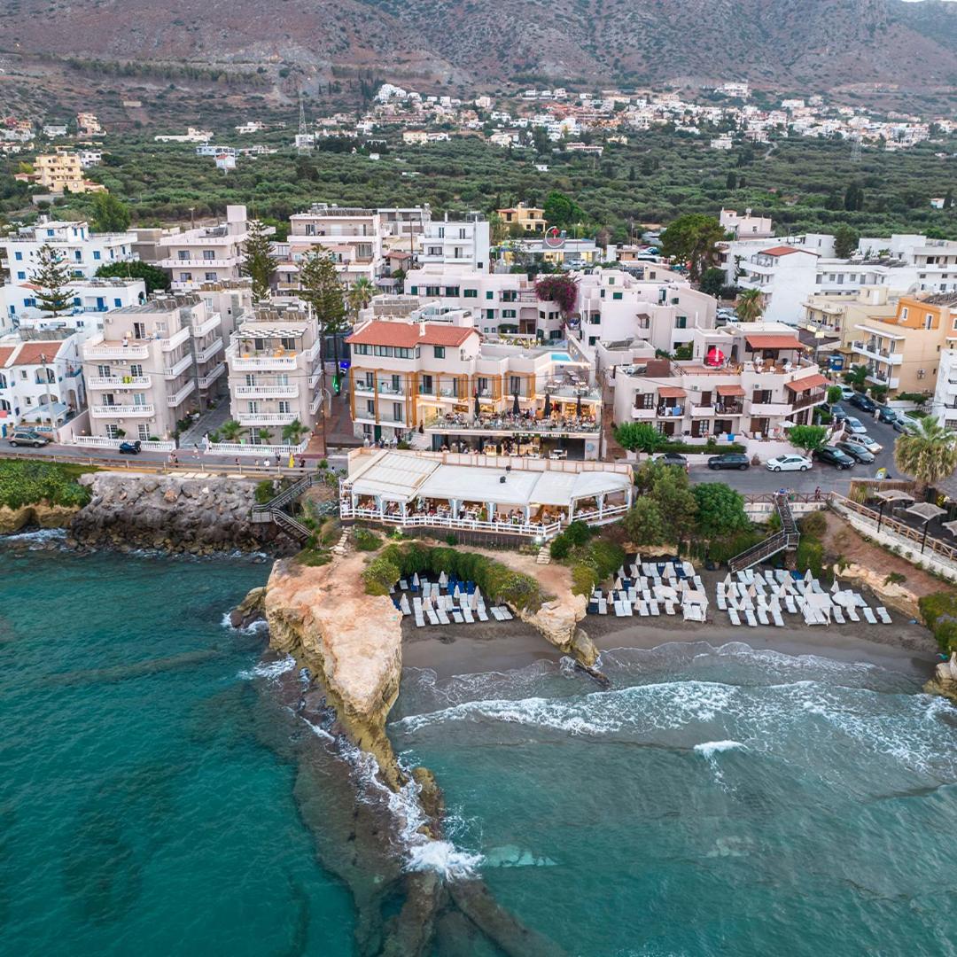 Porto Greco Village panorāmas skats uz viesnīcas kompleksu Hersonisas piekrastē ar pludmali, restorāniem un apkārtējiem kalniem fonā