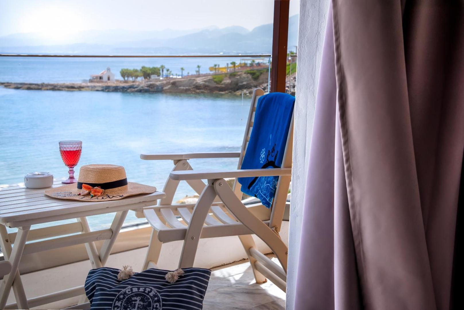 Skats no Porto Greco Village balkona uz jūras līci un piekrasti Hersonisā — balts krēsls, galds un vasaras aksesuāri