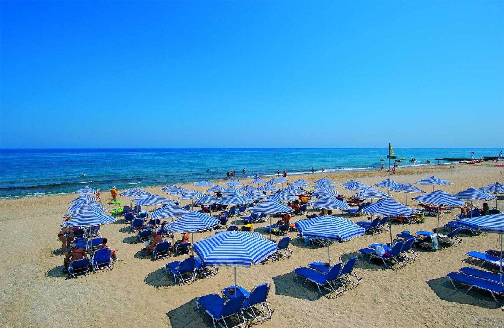 Plaša Eva Bay Hotel smilšu pludmale ar zili svītrainiem saulessargiem un krēsliem pie dzidras jūras Krētā