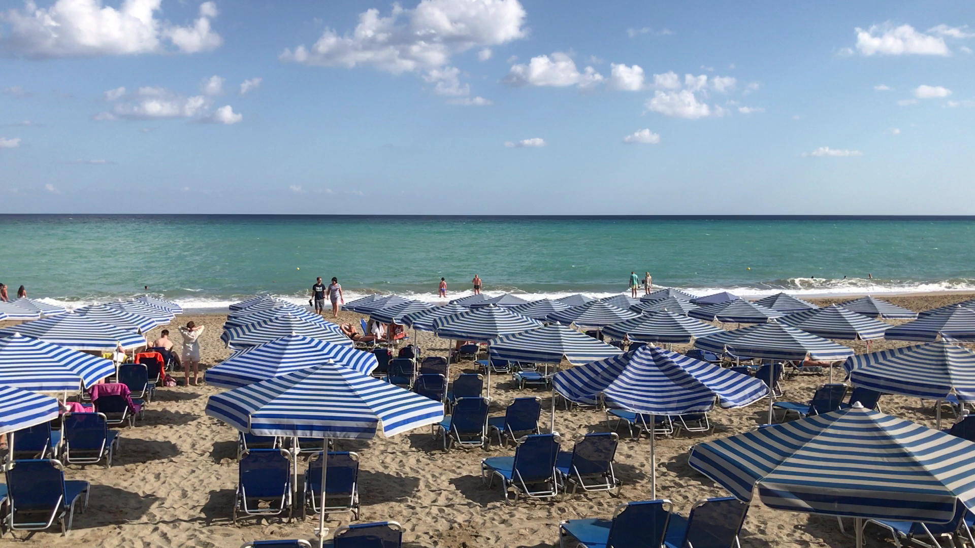 Eva Bay Hotel privātā pludmale Retimno piekrastē Krētā — zili svītraini saulessargi un sauļošanās krēsli pie dzidra tirkīza ūdens