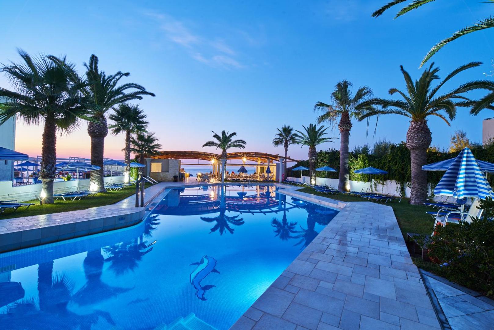 Eva Bay Hotel āra baseins vakarā ar izgaismotām palmām un atpūtas zonu pie jūras Krētā