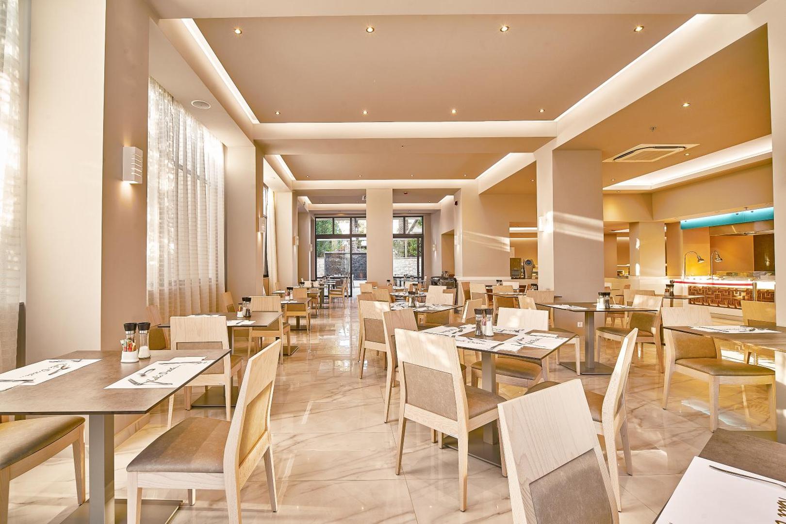 Eva Bay Hotel galvenā restorāna zāle ar elegantiem koka galdiem un krēsliem — gaišs, saulains interjers un marmora flīzes
