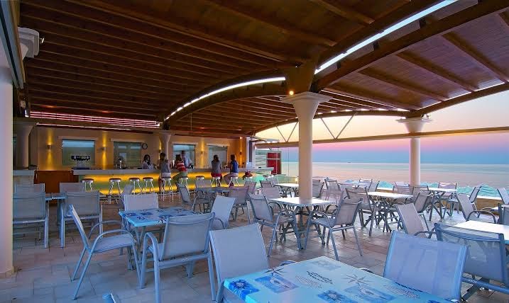 Eva Bay Hotel restorāna terase ar skatu uz Vidusjūru saulrietā — plaša sēdvietu zona zem koka konstrukcijas nojumes