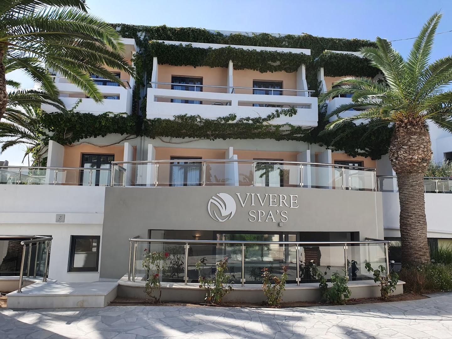 Eva Bay Hotel Vivere Spa ieeja ar palmu koku un modernu fasādi — relaksācijas centrs viesnīcas teritorijā Krētas salā