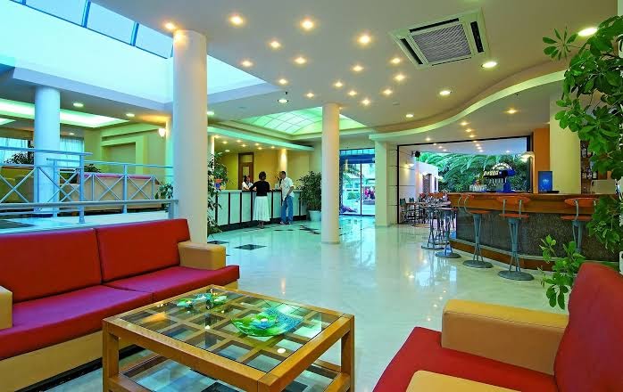 Eva Bay Hotel plašais vestibils ar recepciju, bāru un sarkaniem dīvāniem — moderna un gaiša viesnīcas ieejas zona Krētā