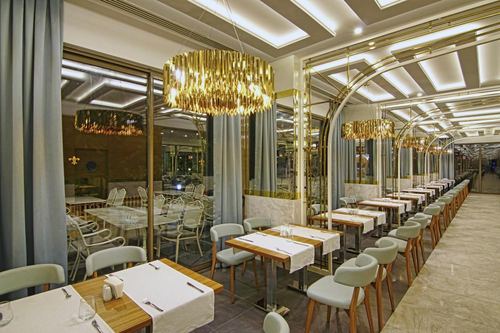 Rai Premium Tekirova à la carte restorāna interjers ar elegantiem galdiem, zelta lustrām un lieliem logiem uz terasi vakara stundās.
