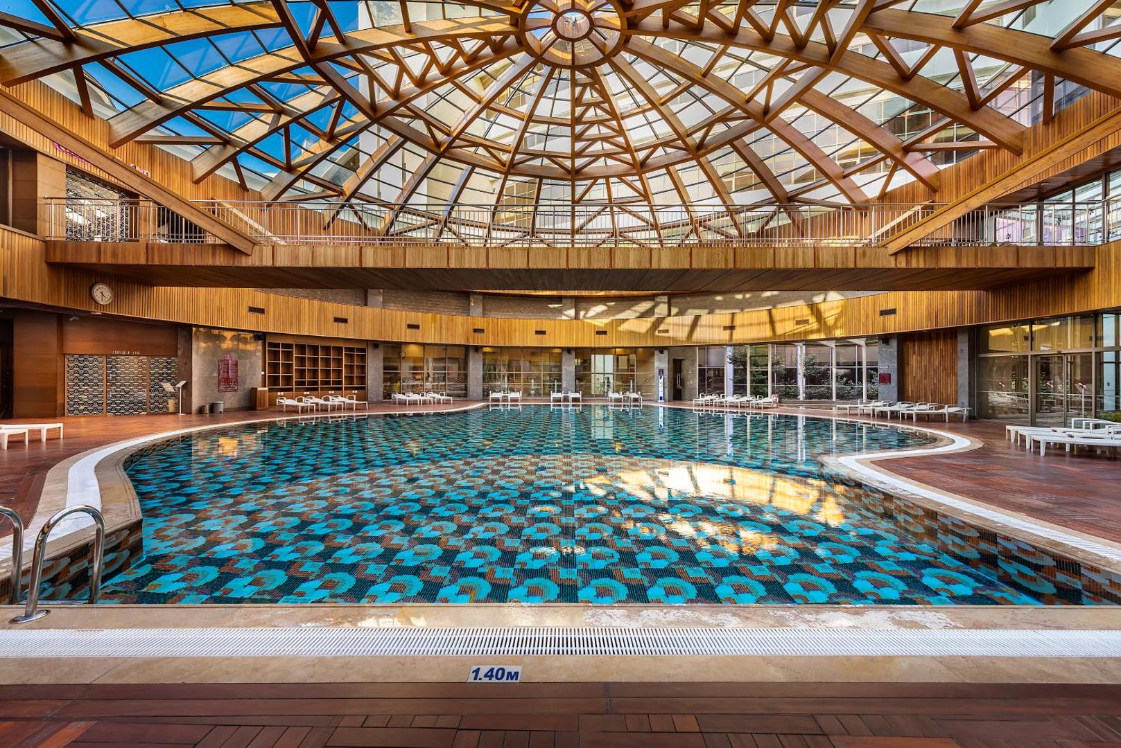 Cornelia Diamond Golf Resort & Spa iekštelpu baseins zem iespaidīga koka un stikla kupola — atpūtas zona ar sauļošanās krēsliem
