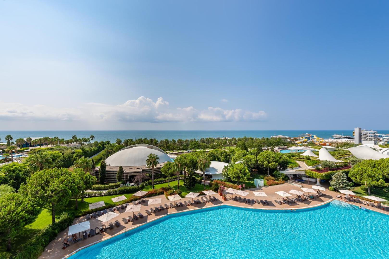 Cornelia Diamond Golf Resort & Spa panorāmas skats uz āra baseinu, dārzu un Vidusjūru Belekā — plaša teritorija ar saulessargiem