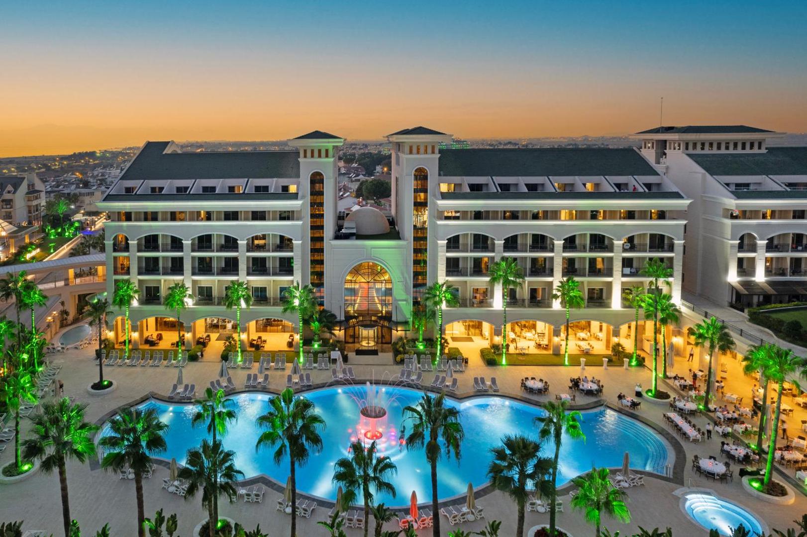Dobedan Exclusive Hotel & Spa galvenā ēka un izgaismots āra baseins saulrietā Belekā — grezna fasāde ar palmām un strūklaku