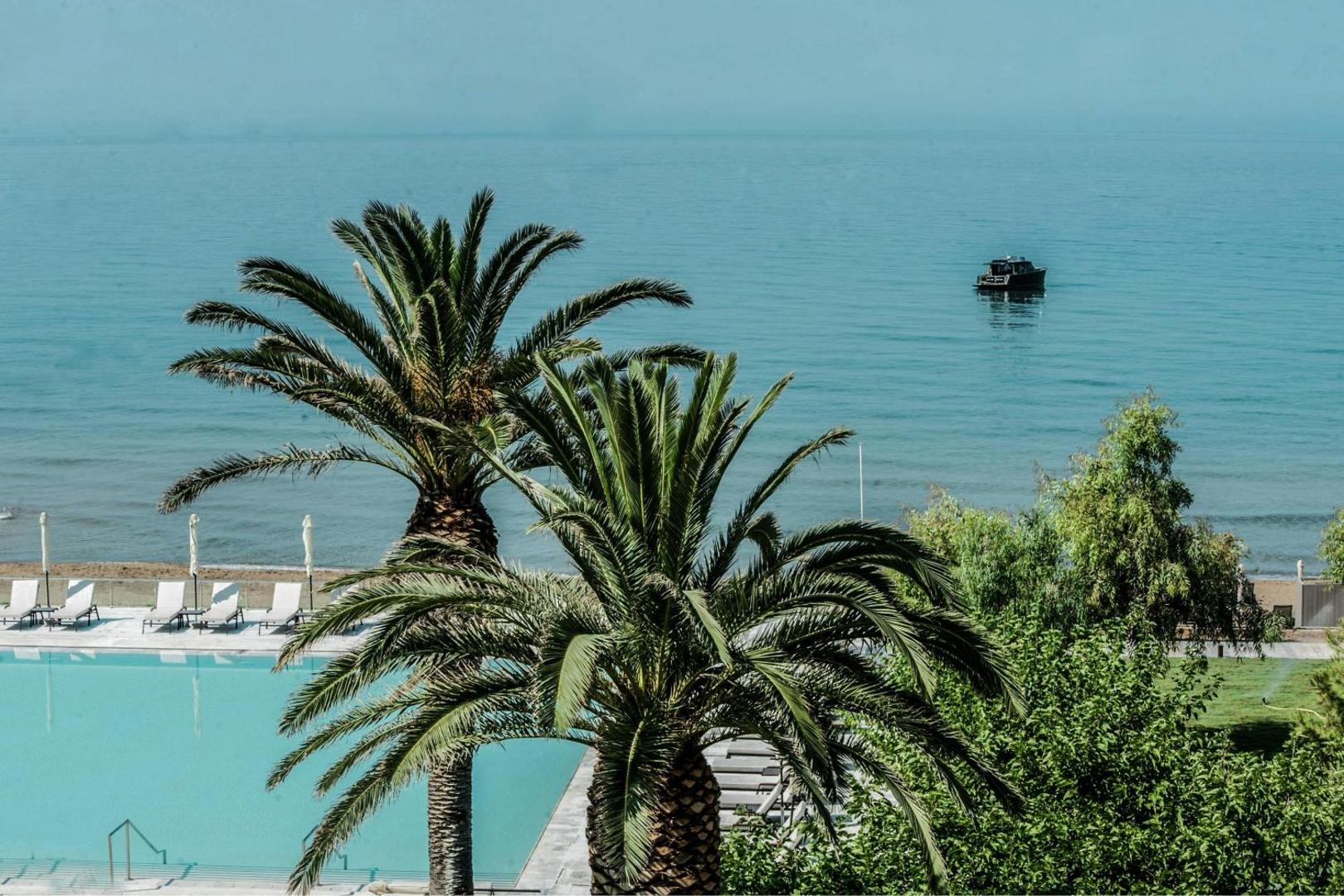 Skats uz Jonijas jūru caur palmu lapām Domes of Corfu viesnīcā, fonā neliela laiva un baseina mala