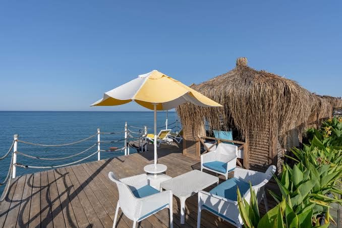 Limak Atlantis De Luxe atpūtas zona uz koka terases ar baltiem krēsliem, dzeltenu saulessargu un bungalo ar niedru jumtu