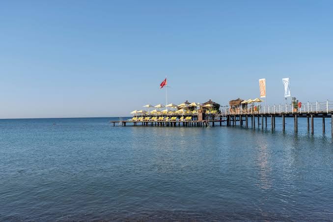 Limak Atlantis De Luxe Hotel & Resort koka piestātne ar sauļošanās krēsliem un saulessargiem Belekas piekrastē virs dzidra jūras ūdens