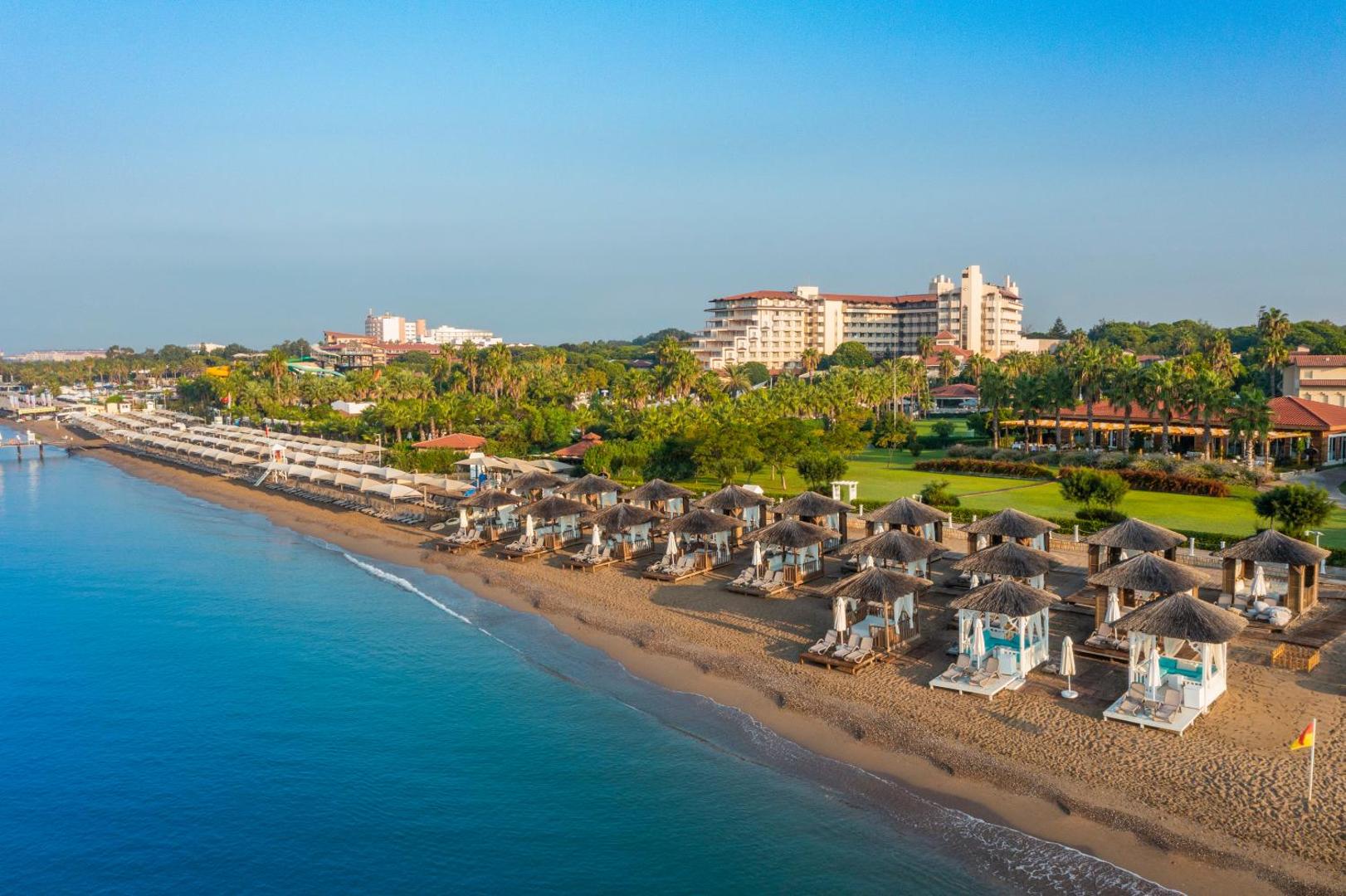 Bellis Deluxe Hotel piekrastes skats saulrietā — pludmales nojumes ar salmu jumtiem un viesnīcas galvenā ēka dārza ieskāvumā