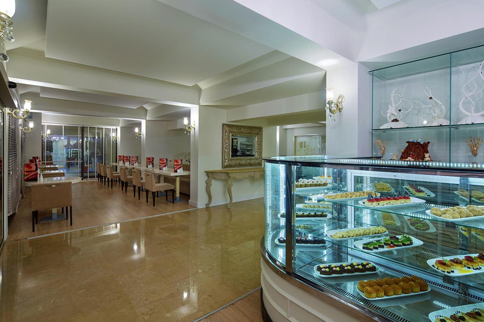 Bellis Deluxe Hotel konditorejas vitrīna ar krāsainiem desertiem un kūkām — gaišs interjers ar galdiņiem viesu uzņemšanai