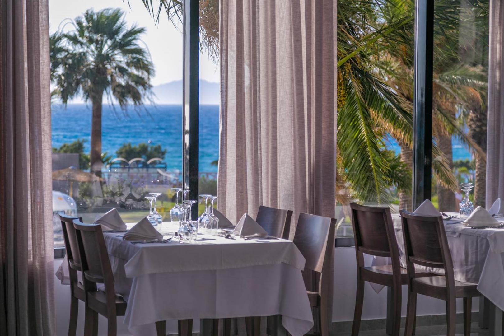 Akti Imperial Deluxe Resort restorāna skats uz jūru Rodas salā — eleganti klāti galdi ar baltiem galdautiem pie lieliem logiem