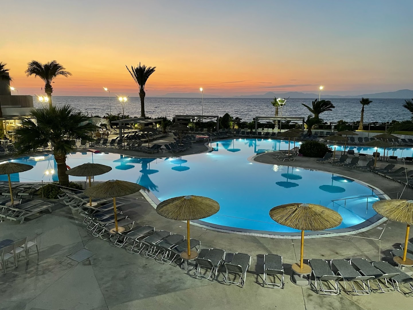 Vakara skats uz Akti Imperial Deluxe Resort & Spa izgaismoto baseinu un saulrietu virs Egejas jūras Rodā