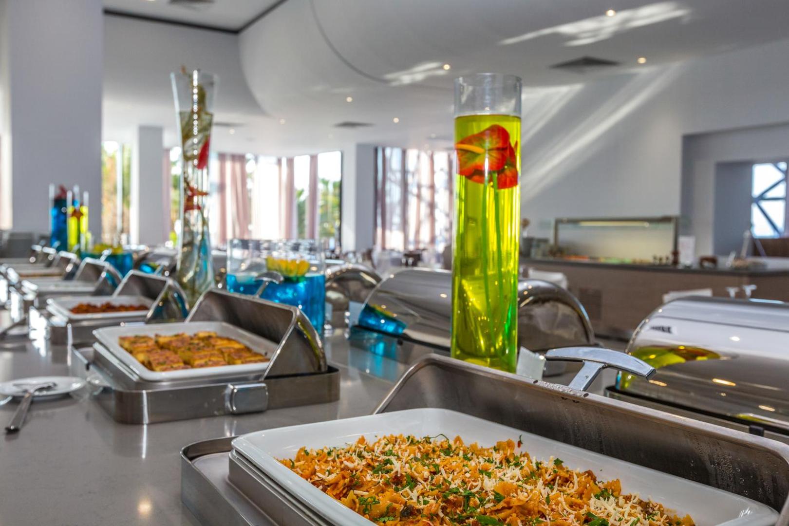 Akti Imperial Deluxe Resort bufetes tipa restorāna līnija ar siltajiem ēdieniem un dekoratīvām vāzēm modernā interjerā