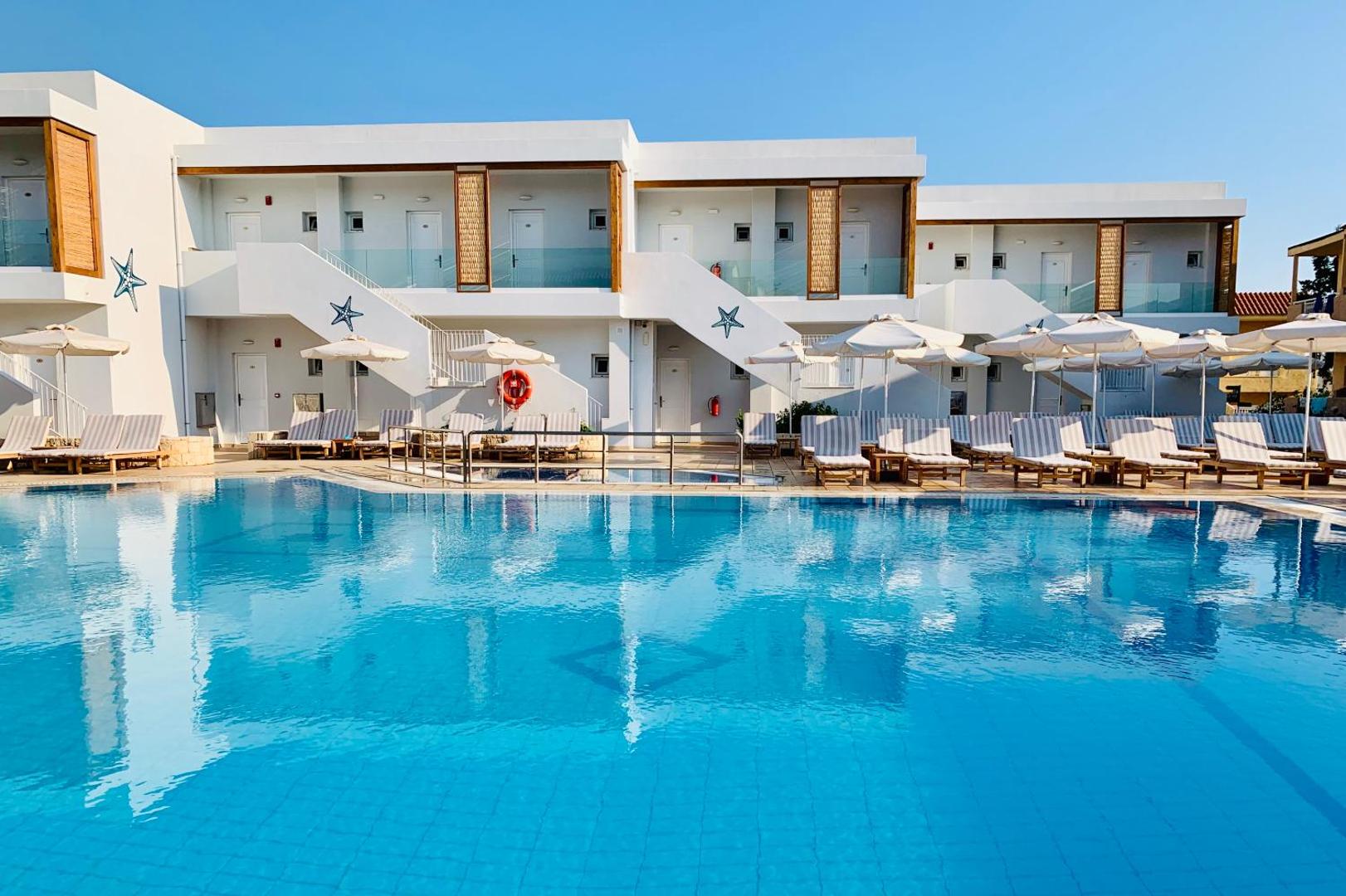 Plašs Aelius Hotel and Spa āra baseins ar saulessargiem un atpūtas krēsliem pie viesnīcas korpusiem saulainā dienā Krētā.