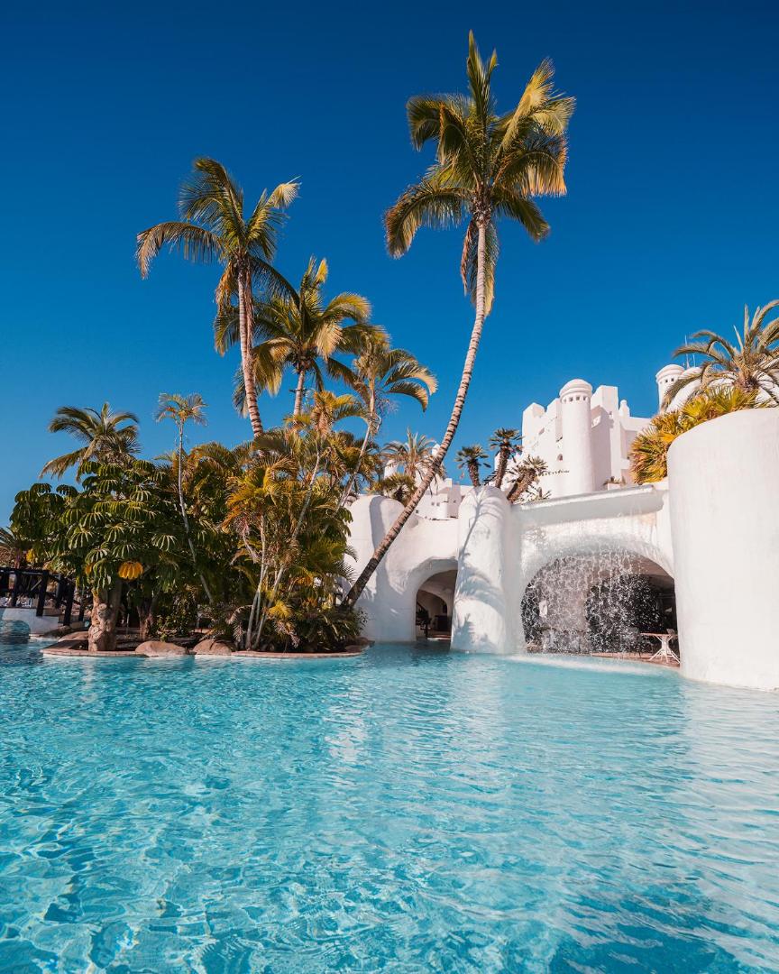 Dreams Jardin Tropical Resort & Spa āra baseins ar kaskādes ūdenskritumu un palmām uz zilu debesu fona