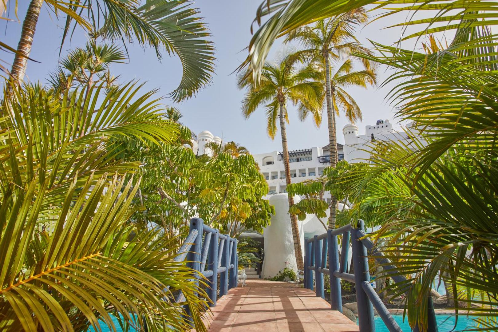 Dreams Jardin Tropical Resort & Spa dārza skats ar koka tiltiņu, palmām un viesnīcas balto fasādi Tenerifē, Kanāriju salās
