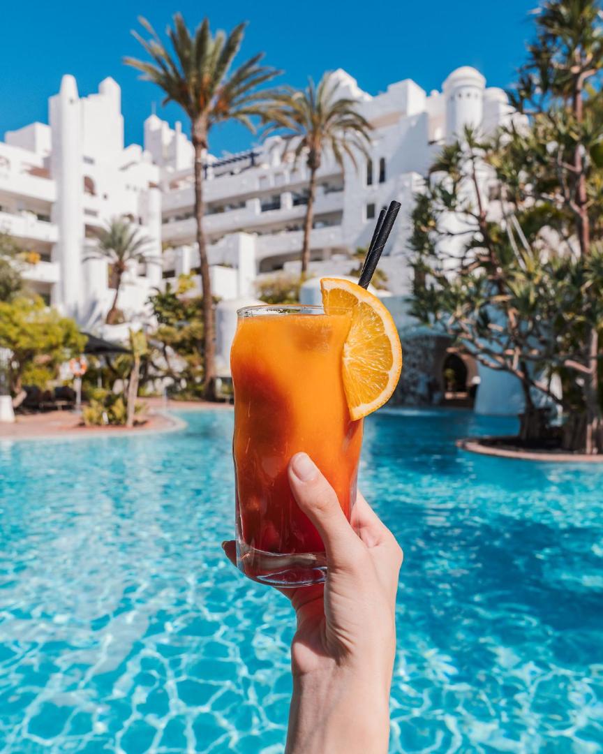 Dreams Jardin Tropical Resort & Spa baseina zona ar zilu ūdeni, palmu koku fonu un roku, kas tur krāsainu augļu kokteili