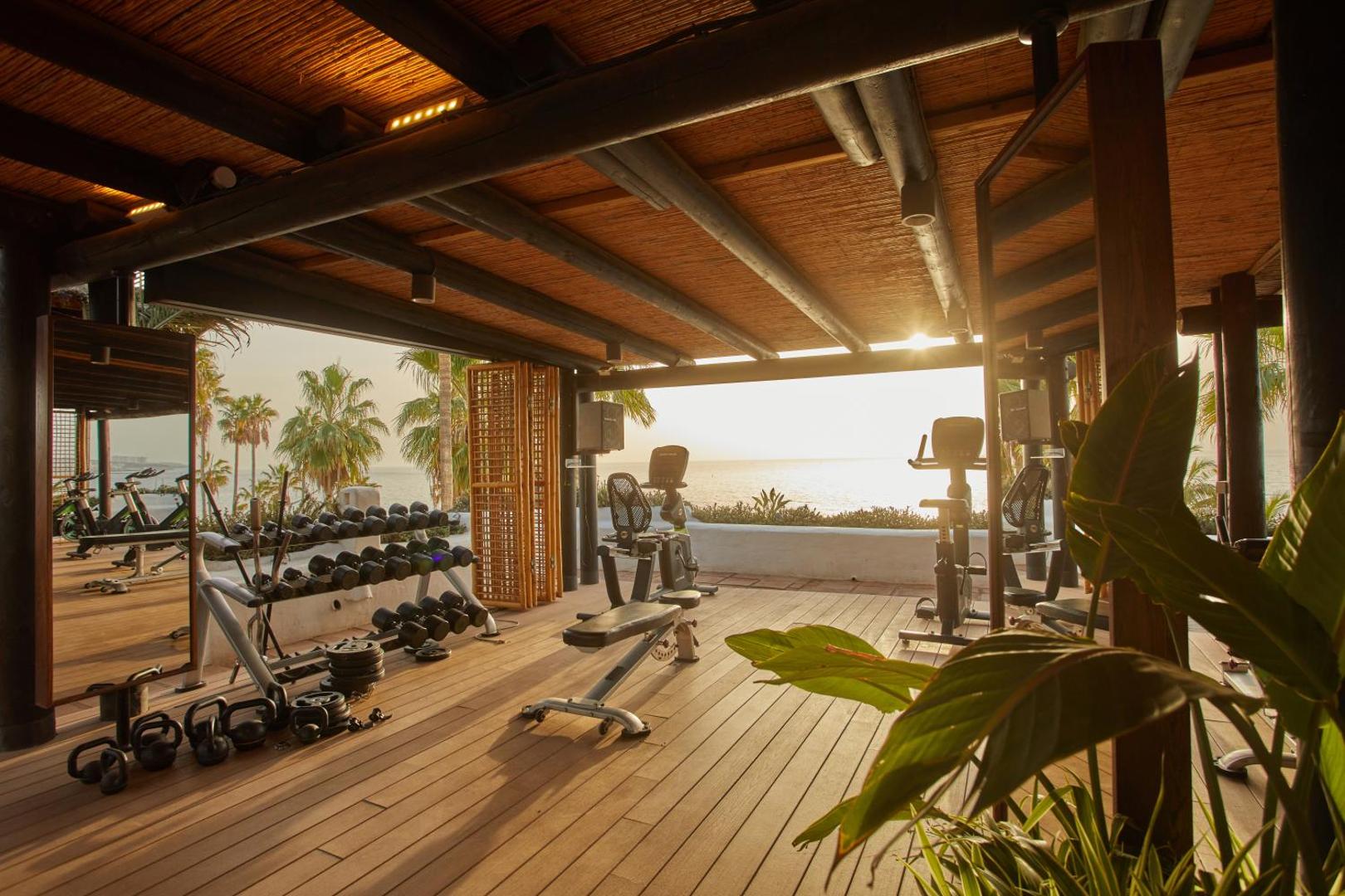 Dreams Jardin Tropical Resort & Spa brīvdabas fitness centrs Tenerifē — trenažieri un svari uz koka terases saulrieta laikā