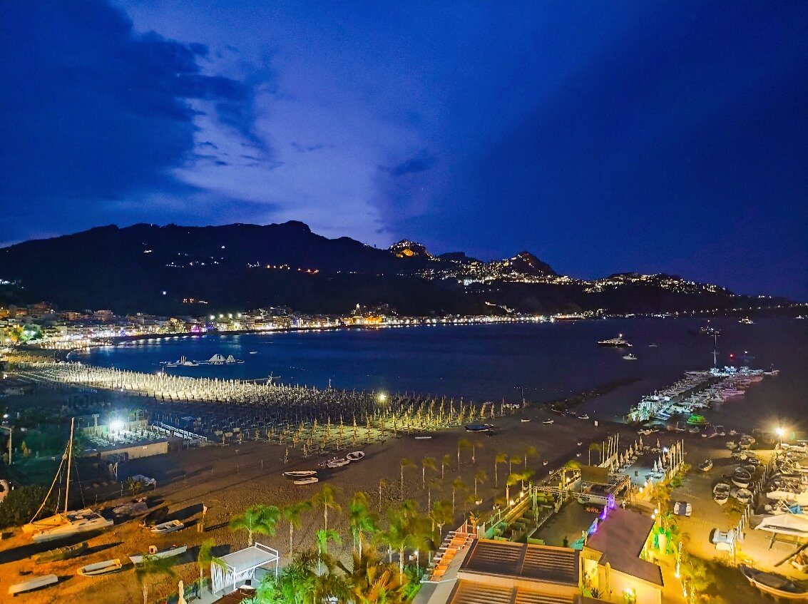 Panoramic viesnīcas skats naktī uz izgaismoto Giardini Naxos līci Sicīlijā — pilsētas gaismas atspīd ūdenī, pludmales josla un jahtu piestātne.