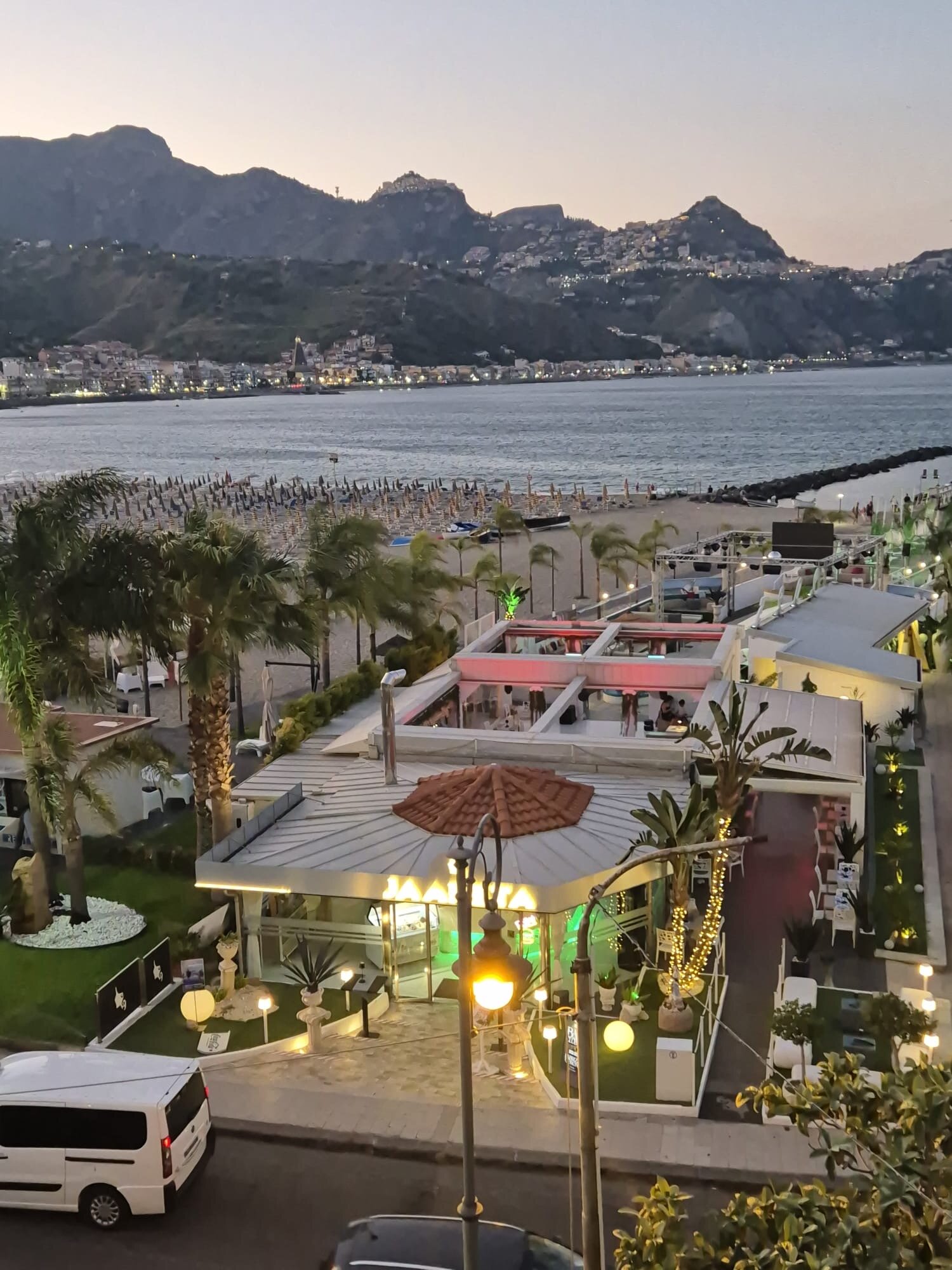 Skats no Hotel Panoramic uz pludmales zonu un piekrastes bāru vakarā Giardini Naxos, Sicīlijā ar kalnu fonu