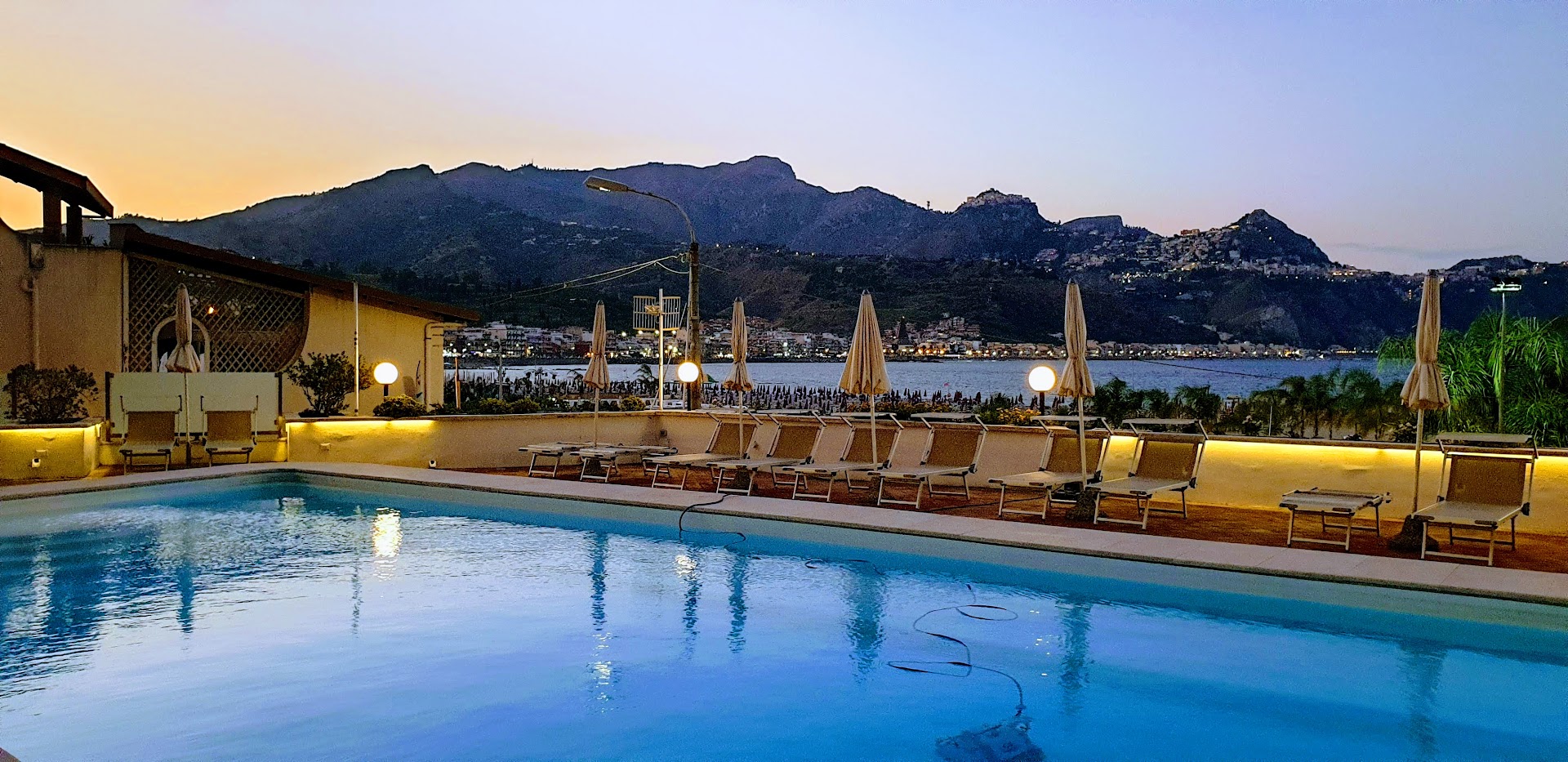 Hotel Panoramic āra baseins saulrieta laikā Giardini Naxos, Sicīlijā — skats uz izgaismotu terasi, sauļošanās krēsliem un kalnu siluetu fonā