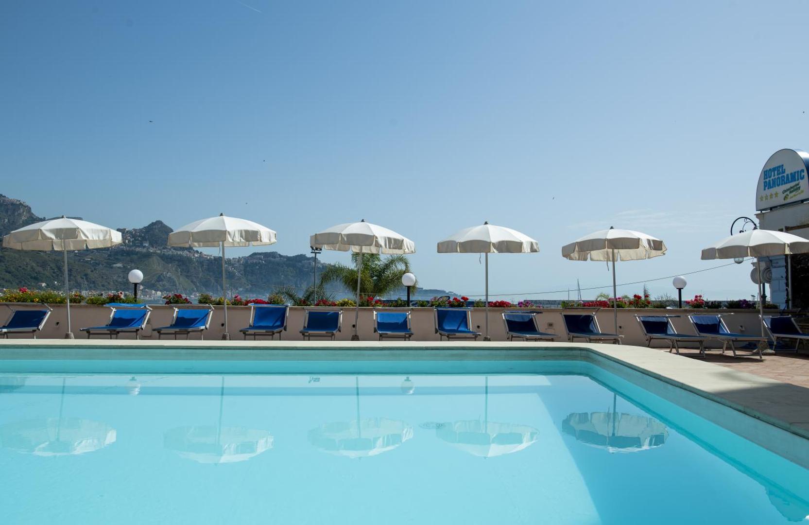 Hotel Panoramic baseina mala ar baltiem saulessargiem un zilām atpūtas gultām Giardini Naxos, Sicīlijā — fonā redzami kalni un jūra