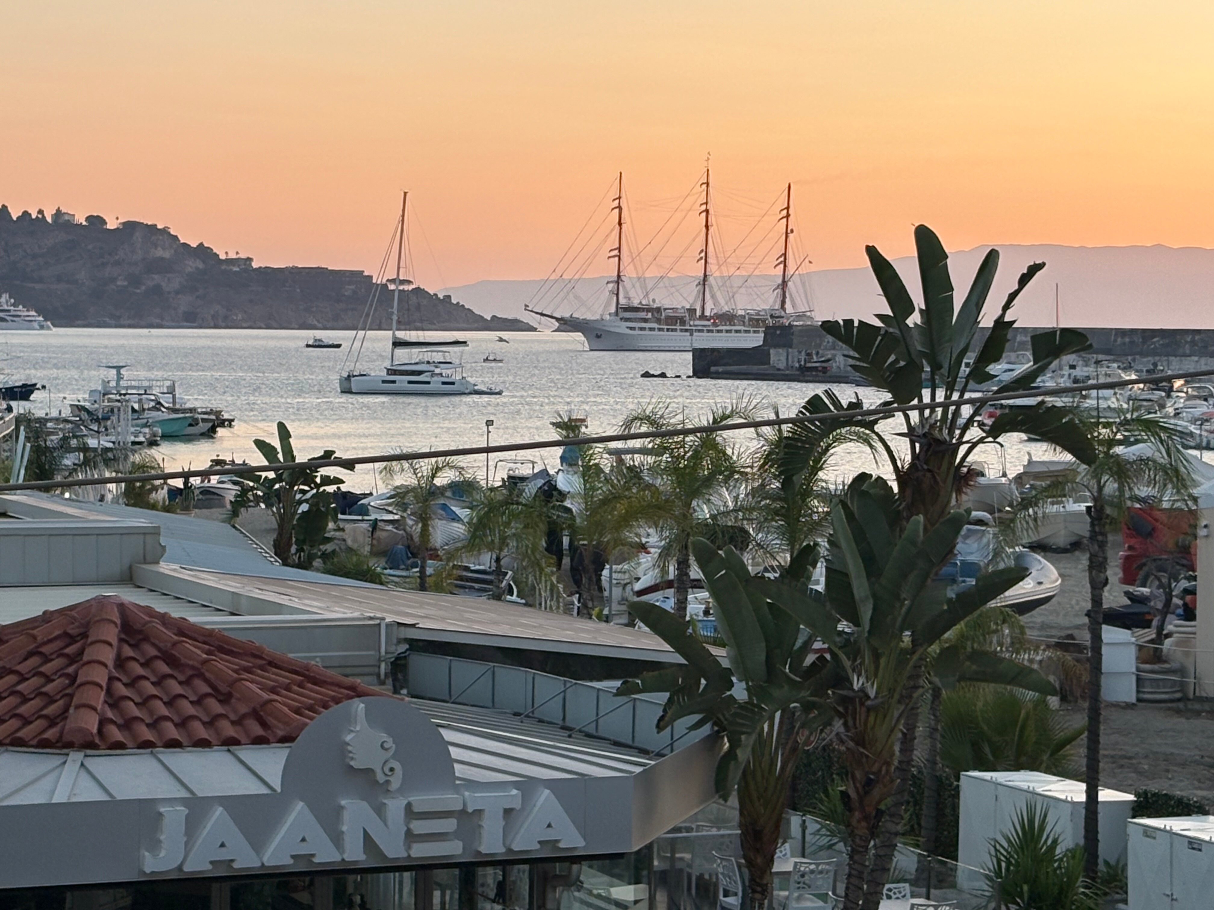 Skats no viesnīcas Panoramic terases uz Giardini Naxos līci saulrietā ar jahtām un burinieku piekrastē, Sicīlijā.