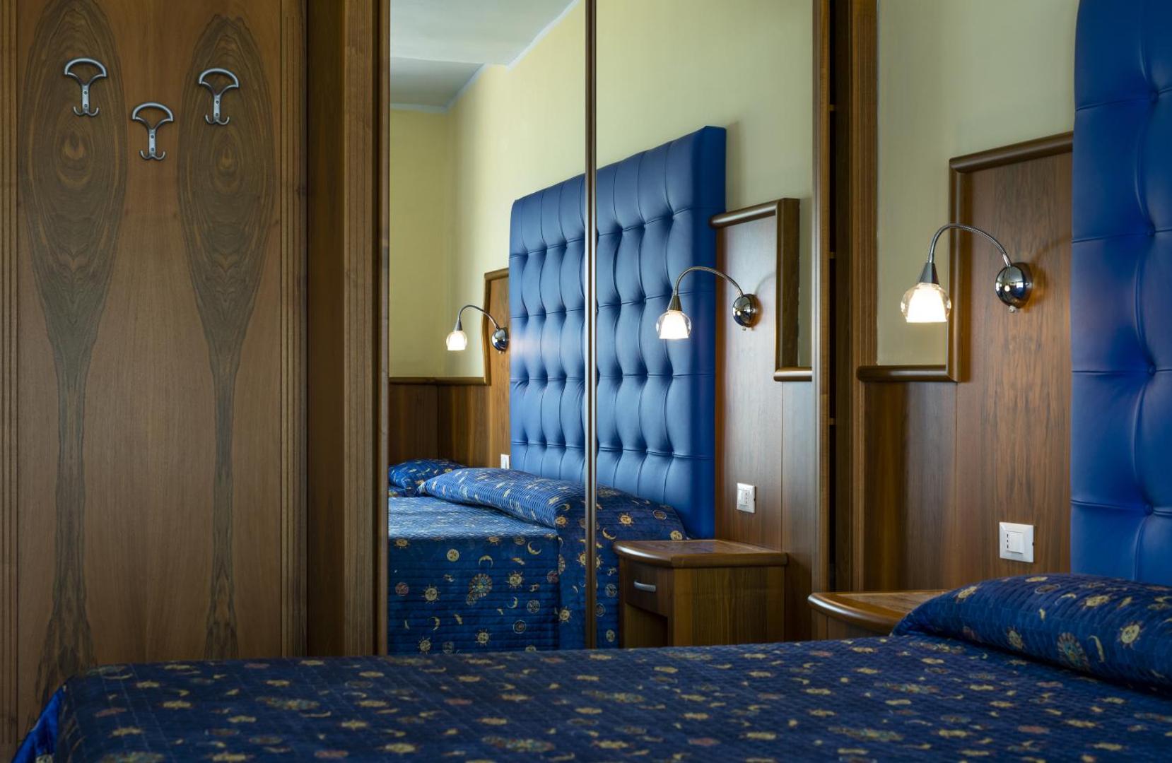 Panoramic viesnīcas numura interjers ar zilu galvgali un koka apdari — spoguļi un mājīgs apgaismojums klasiskā stilā.