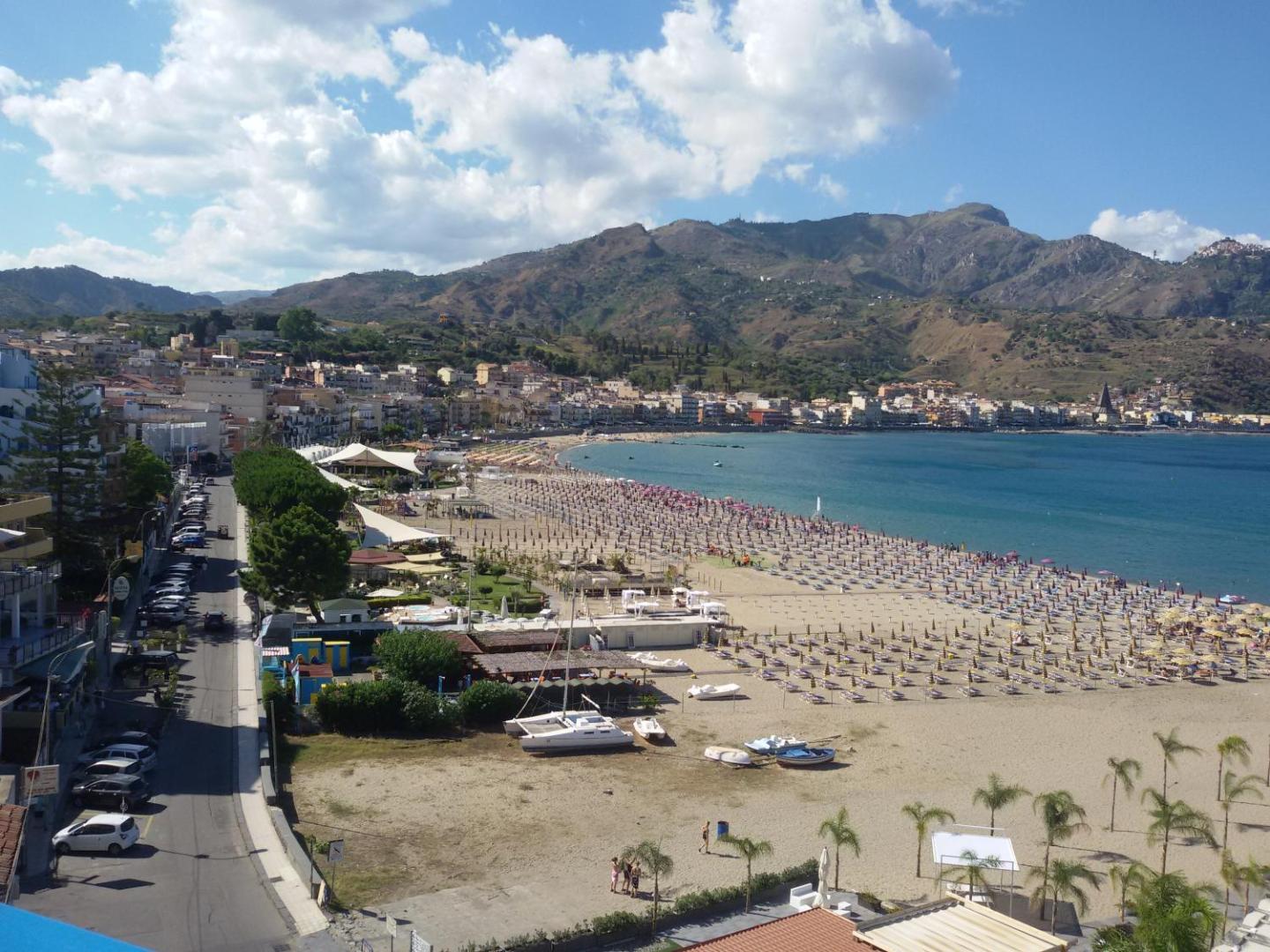Skats no Panoramic viesnīcas uz Giardini Naxos pludmali Sicīlijā — plaša smilšu piekraste ar saulessargiem, pilsētas panorāmu un kalniem fonā.
