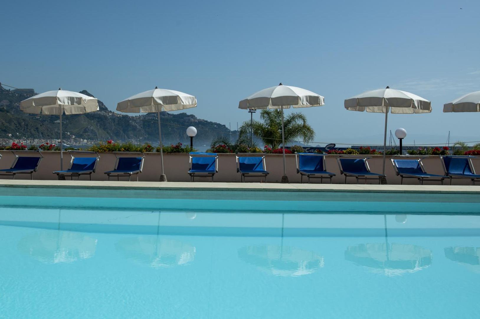 Tuvplāns Hotel Panoramic baseinam ar atspulgiem un baltiem saulessargiem Giardini Naxos piekrastē Sicīlijā — fonā kalnaina ainava