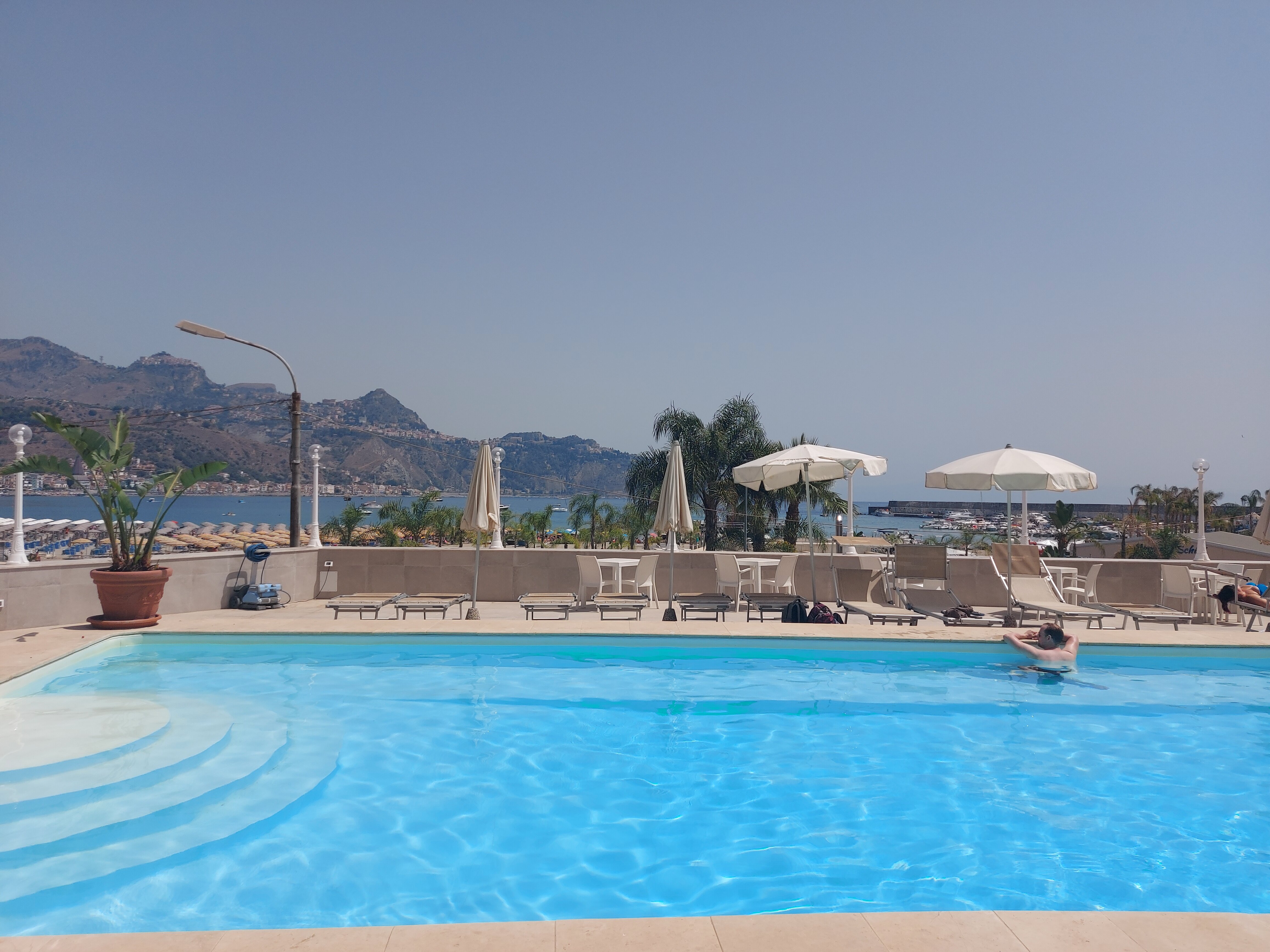 Hotel Panoramic baseins ar skatu uz jūru un Giardini Naxos piekrasti Sicīlijā — gaiši zils ūdens, saulessargi un atpūtas zona