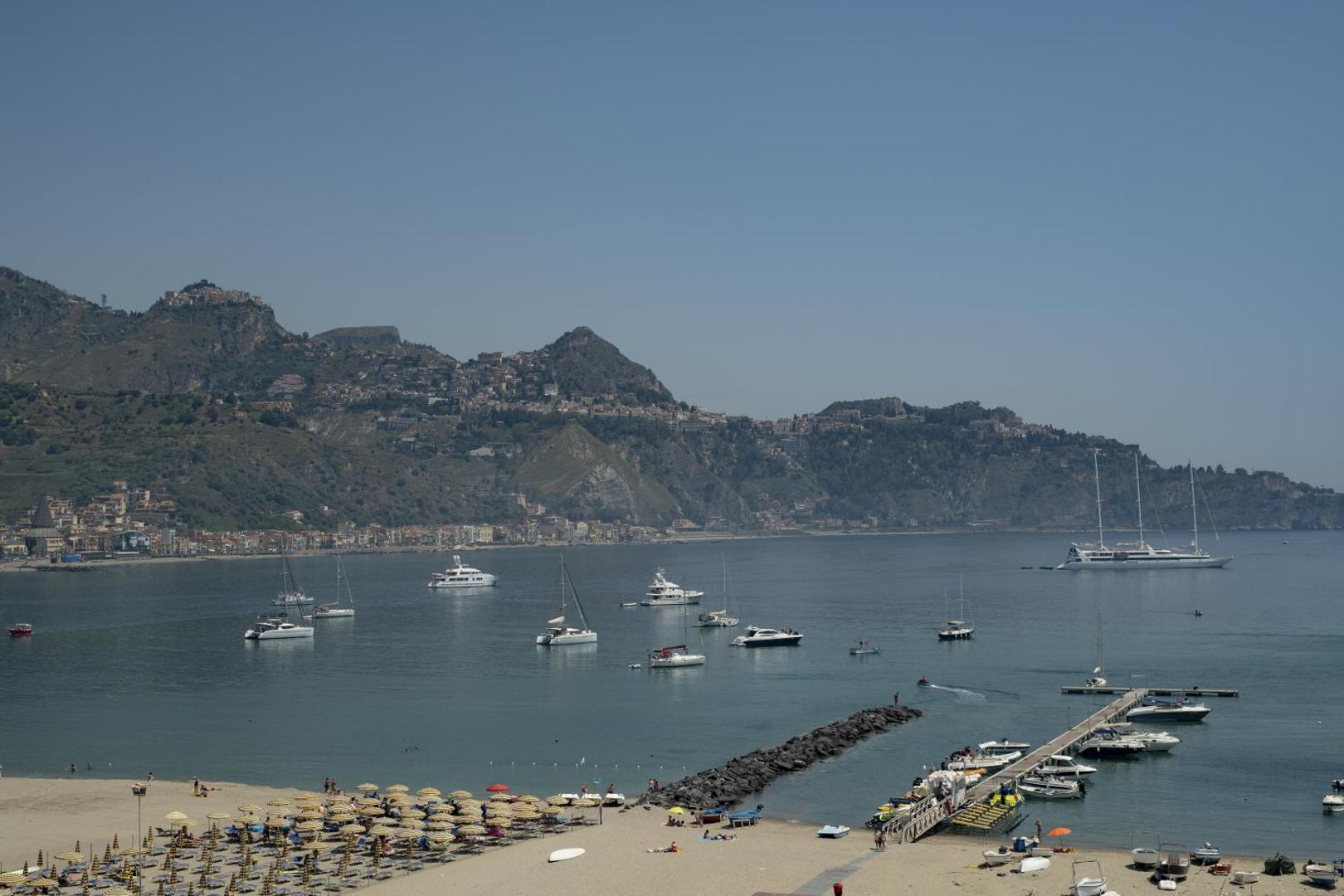 Skats no Panoramic viesnīcas uz Jonijas jūru Sicīlijā — jahtas līcī, pludmales zona ar saulessargiem un Taormina kalnu virotnes.
