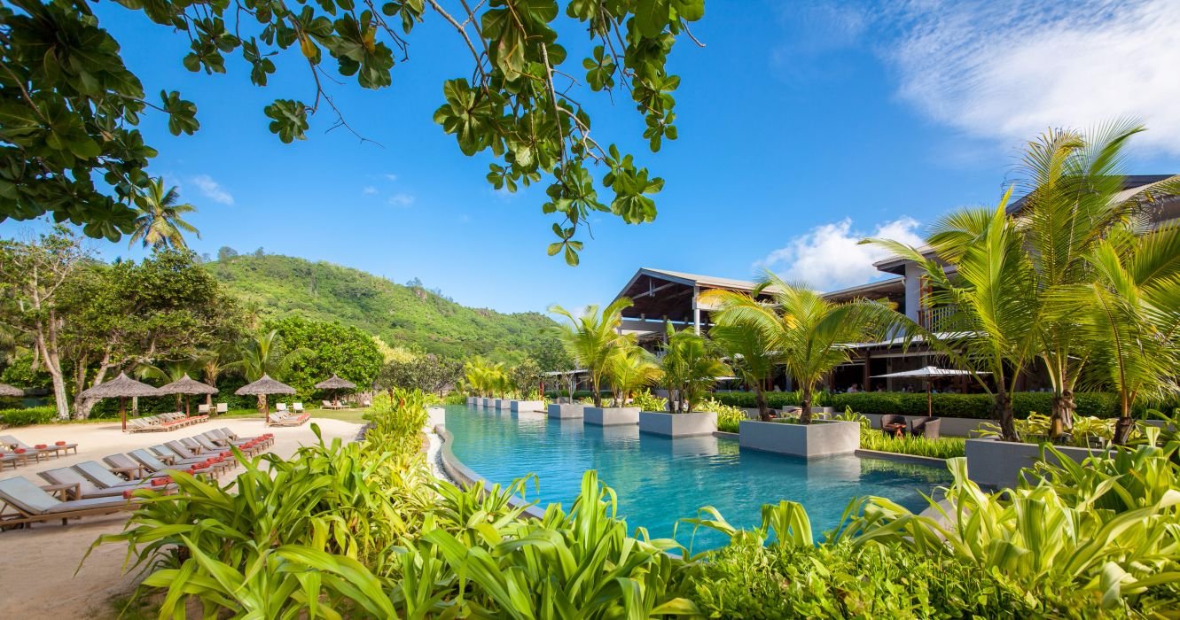 Kempinski Seychelles Resort plašais āra baseins ar palmu saliņām un kalnu ainavu fonā.