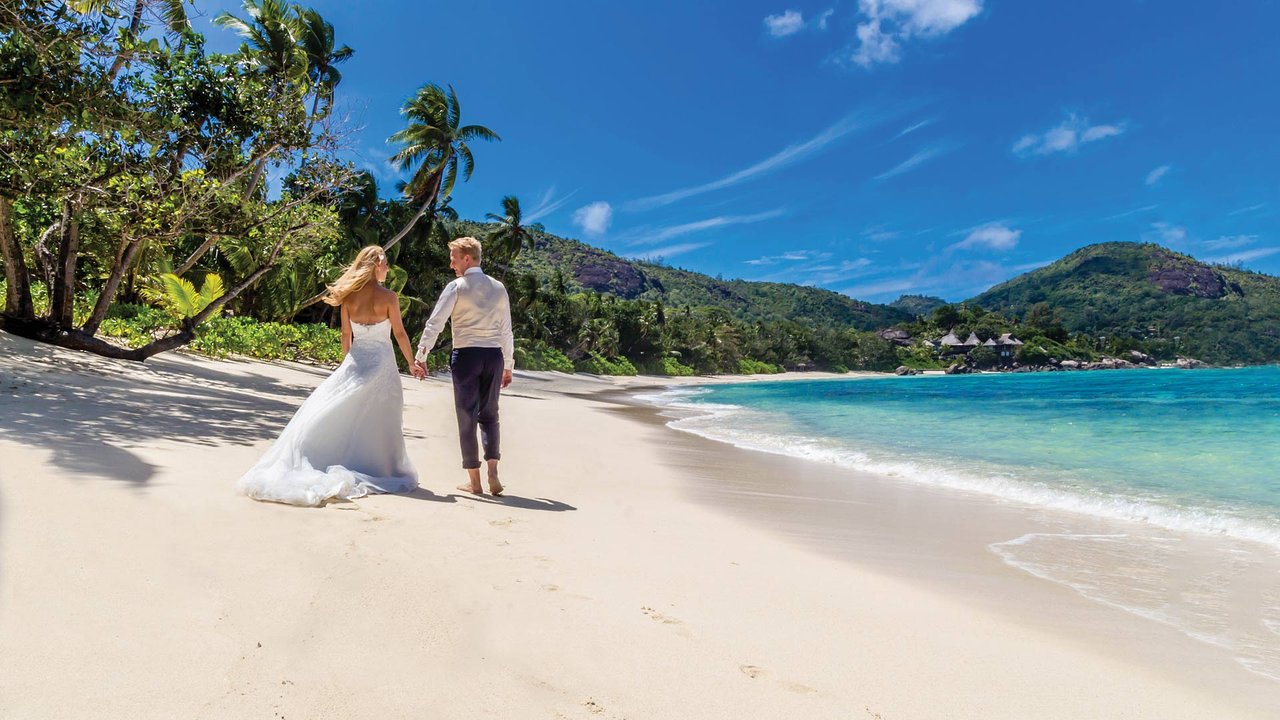 Jaunlaulātie pastaigājas pa balto smilšu pludmali Kempinski Seychelles Resort kāzu ceremonijas laikā.